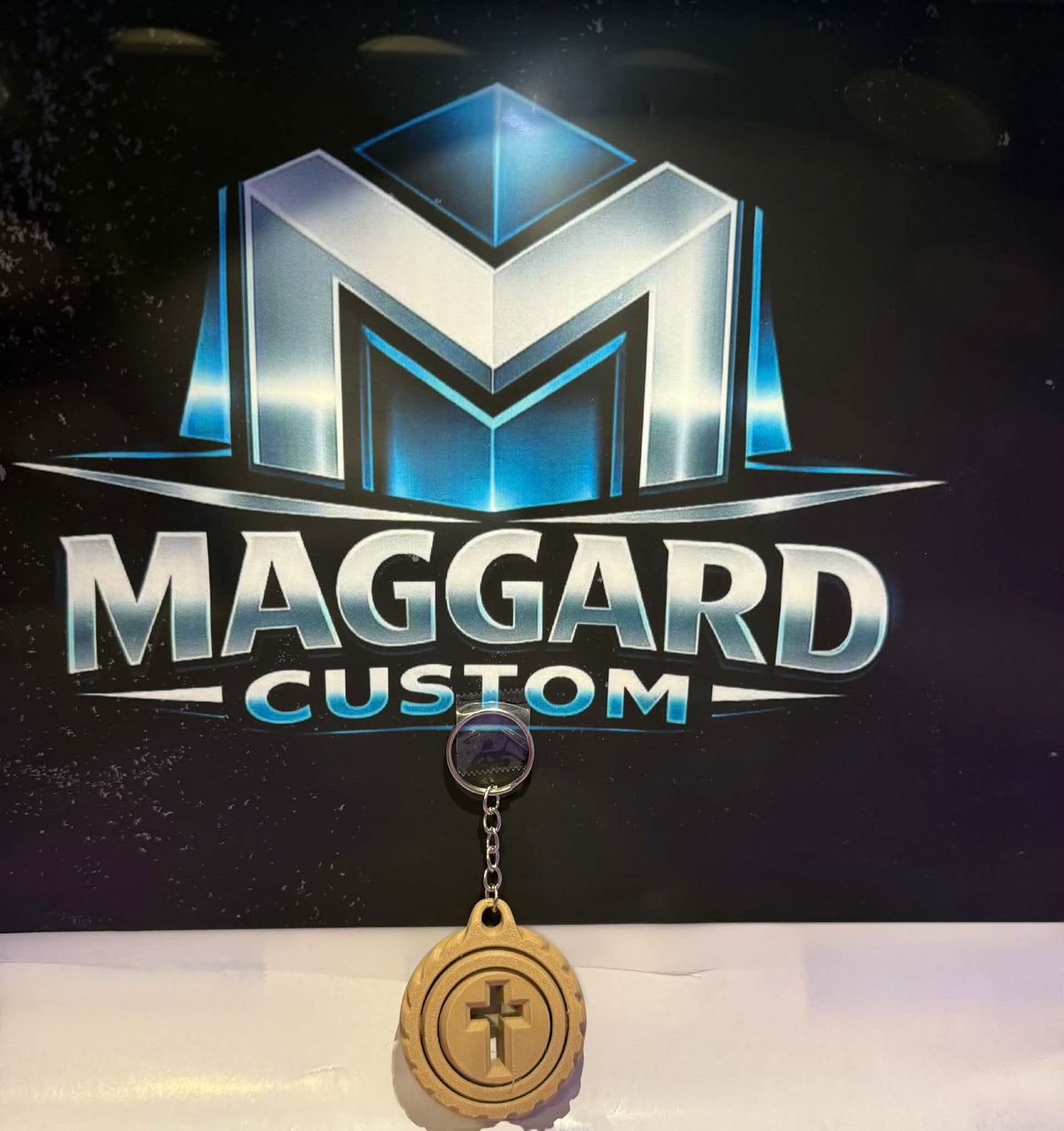 3D printed natural wood-tone cross swirl keychain with raised cross center and decorative ring border with metal keyring displayed in front of Maggard Custom logo