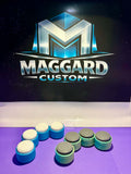 3D printed two-tone drum pad button fidget clickers in blue with white and gray tops displayed in front of Maggard Custom logo
