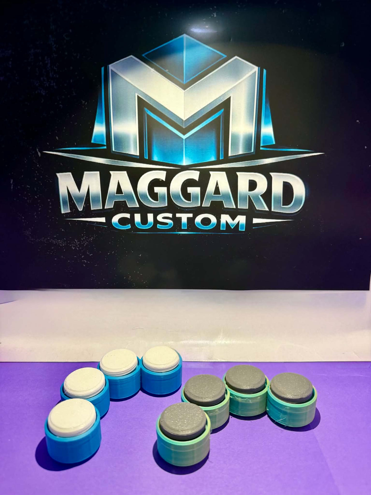 3D printed two-tone drum pad button fidget clickers in blue with white and gray tops displayed in front of Maggard Custom logo