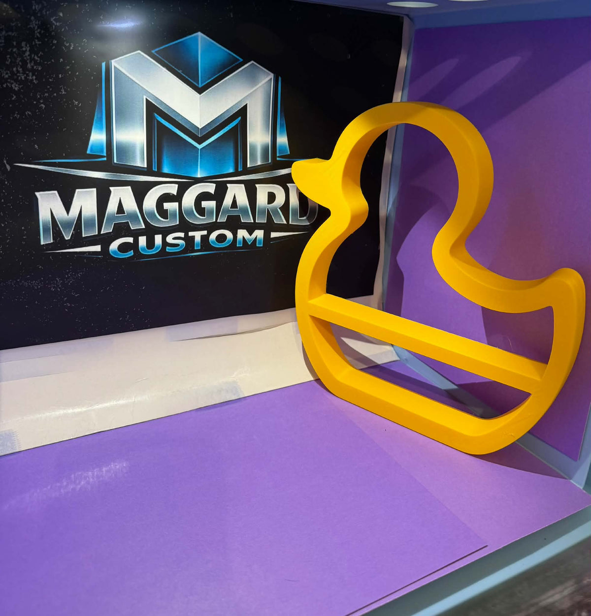 3D printed yellow rubber duck silhouette display shelf with open frame design and built-in ledge displayed in front of Maggard Custom logo