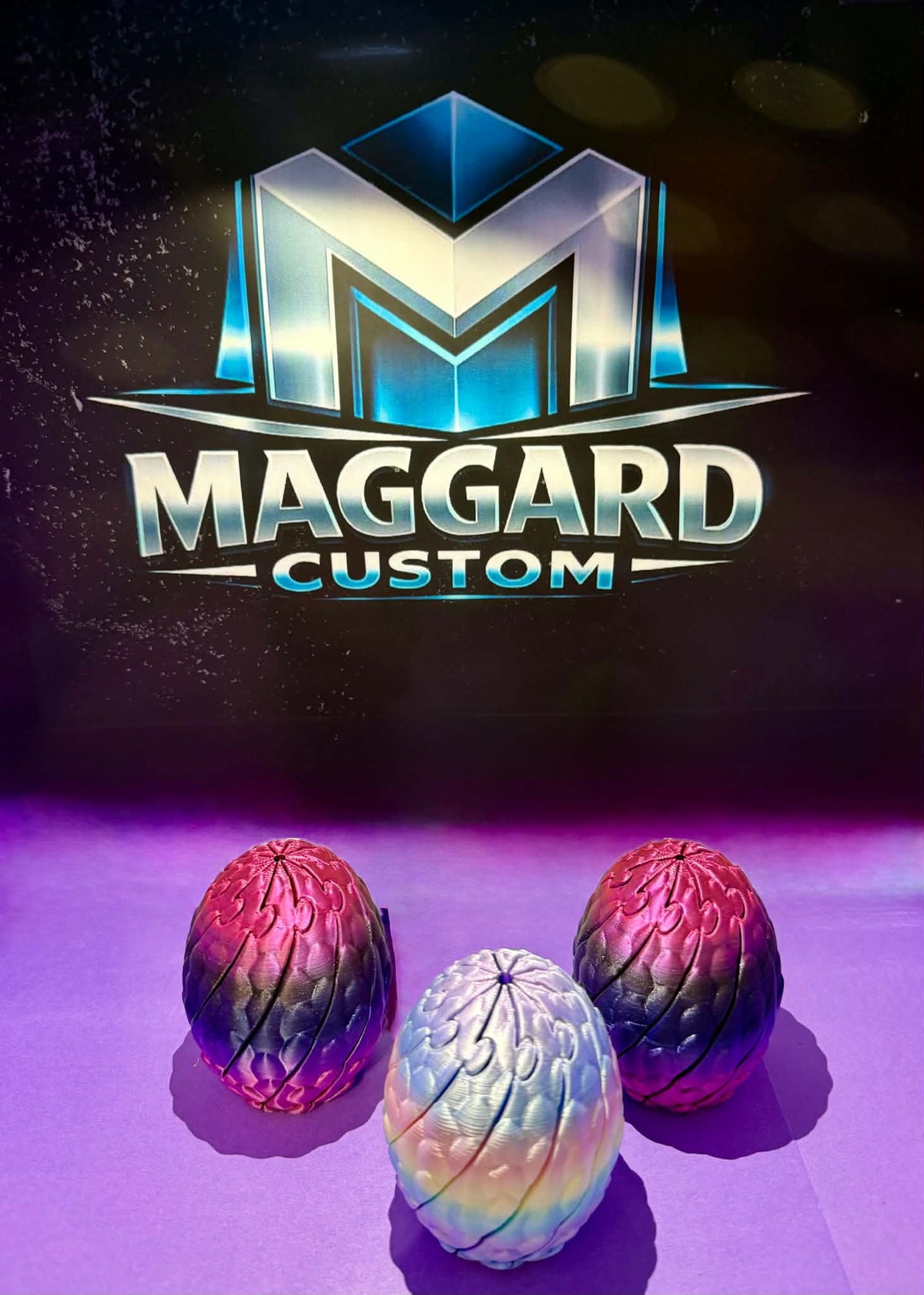 3D printed dragon egg figurines with detailed scale texture in red-purple gradient and iridescent pearl finishes displayed in front of Maggard Custom logo