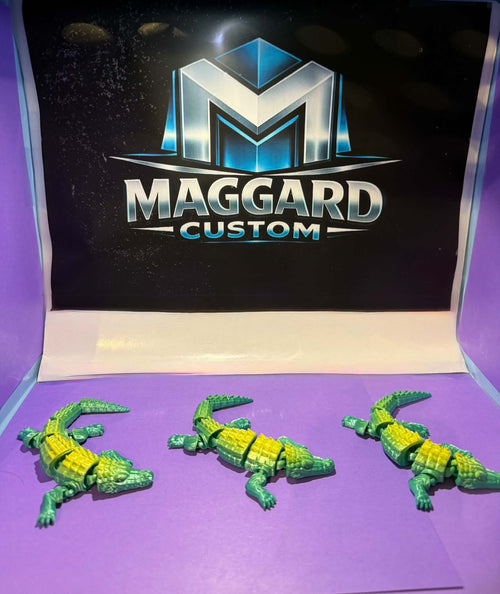 3D printed armored alligator figurines in green and yellow two-tone finish with detailed scale plates displayed in front of Maggard Custom logo