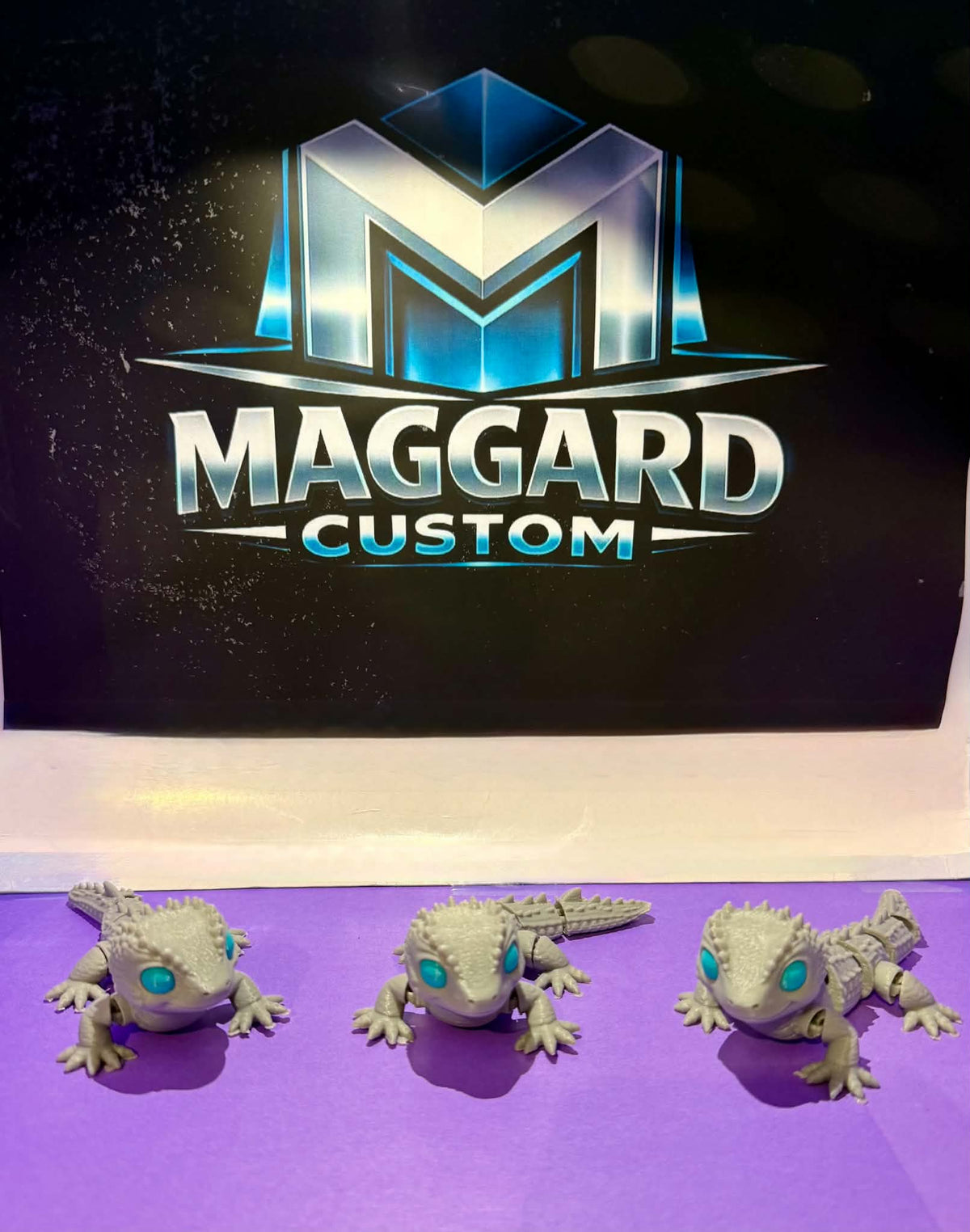3D printed articulated baby dragon gecko figurines in stone gray with teal gemstone eyes shown in three different poses displayed in front of Maggard Custom logo