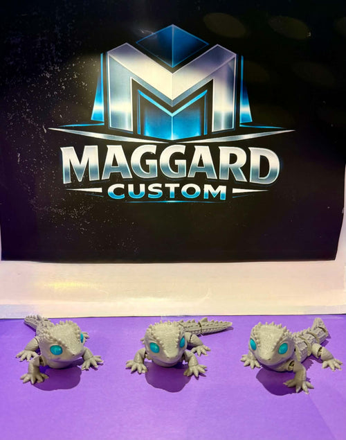 3D printed articulated baby dragon gecko figurines in stone gray with teal gemstone eyes shown in three different poses displayed in front of Maggard Custom logo
