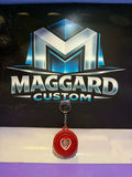 3D printed red heart swirl fidget keychain with cut-out heart center and concentric ring texture with metal keyring displayed in front of Maggard Custom logo