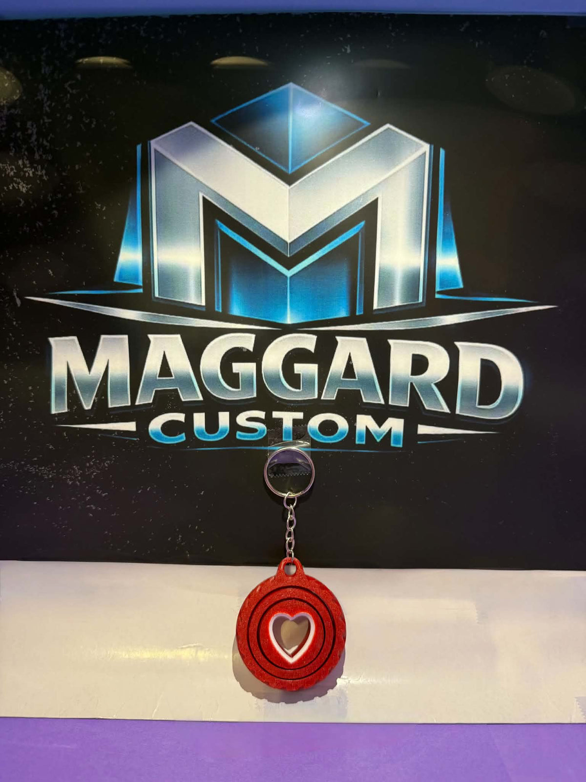 3D printed red heart swirl fidget keychain with cut-out heart center and concentric ring texture with metal keyring displayed in front of Maggard Custom logo