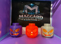3D printed block toy head jack-o-lantern candle holders in purple, orange, and gray glowing with warm light through carved faces displayed in front of Maggard Custom logo