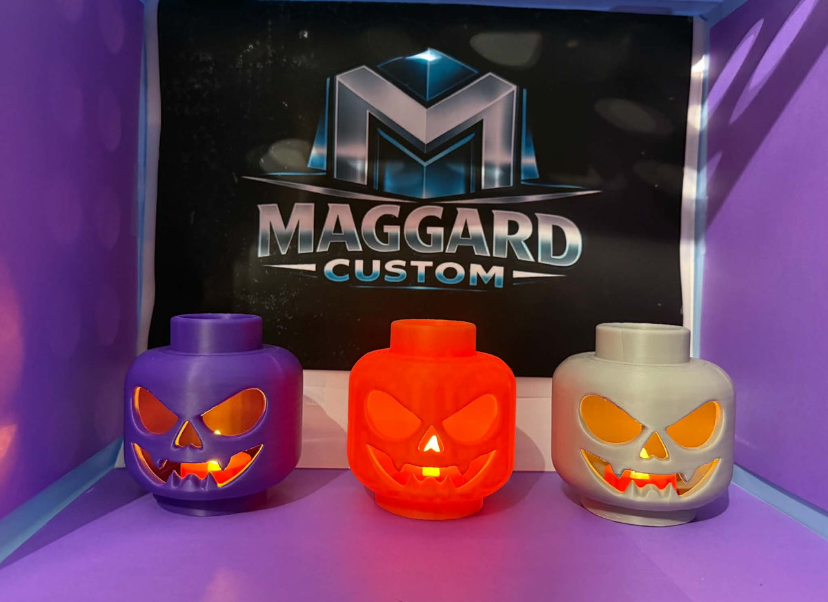 3D printed block toy head jack-o-lantern candle holders in purple, orange, and gray glowing with warm light through carved faces displayed in front of Maggard Custom logo