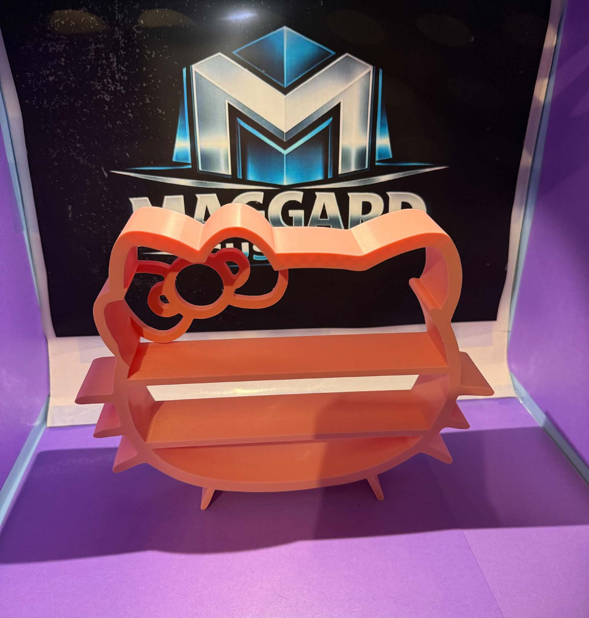 3D printed coral orange cute bow display shelf with scalloped top rail, decorative bow accent, and two display tiers displayed in front of Maggard Custom logo