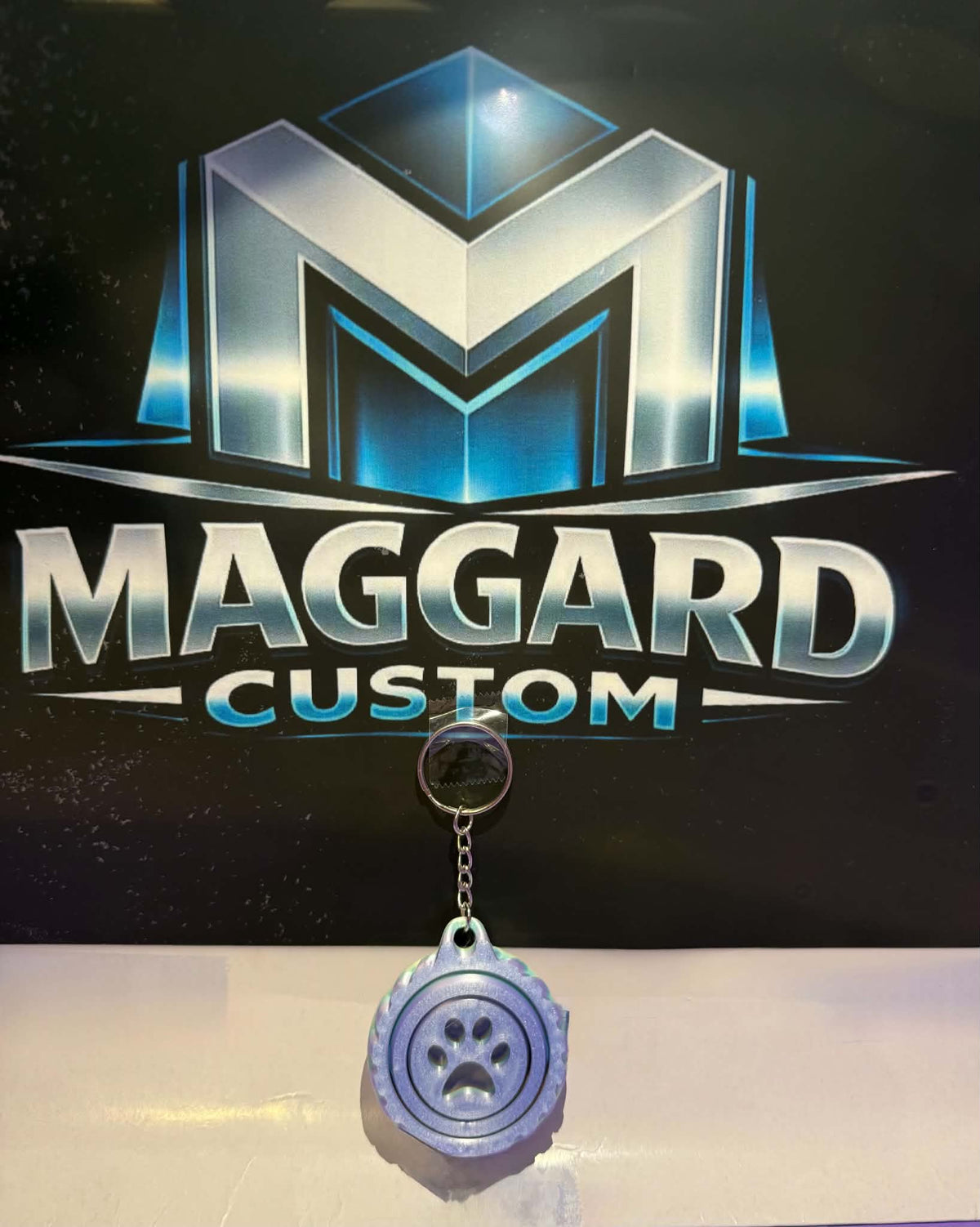 3D printed lavender blue paw print swirl keychain with raised paw center and gear-style textured border with metal keyring displayed in front of Maggard Custom logo