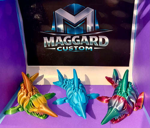 3D printed armored dragon shark figurines in gold-green gradient, solid teal, and red-silver metallic gradient finishes displayed in front of Maggard Custom logo