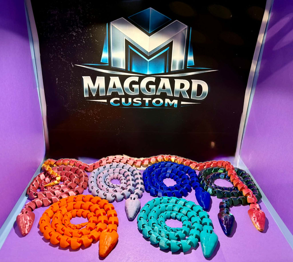 3D printed articulated flexi snake figurines in orange, red, gray, blue, teal, dark green, and purple coiled together displayed in front of Maggard Custom logo
