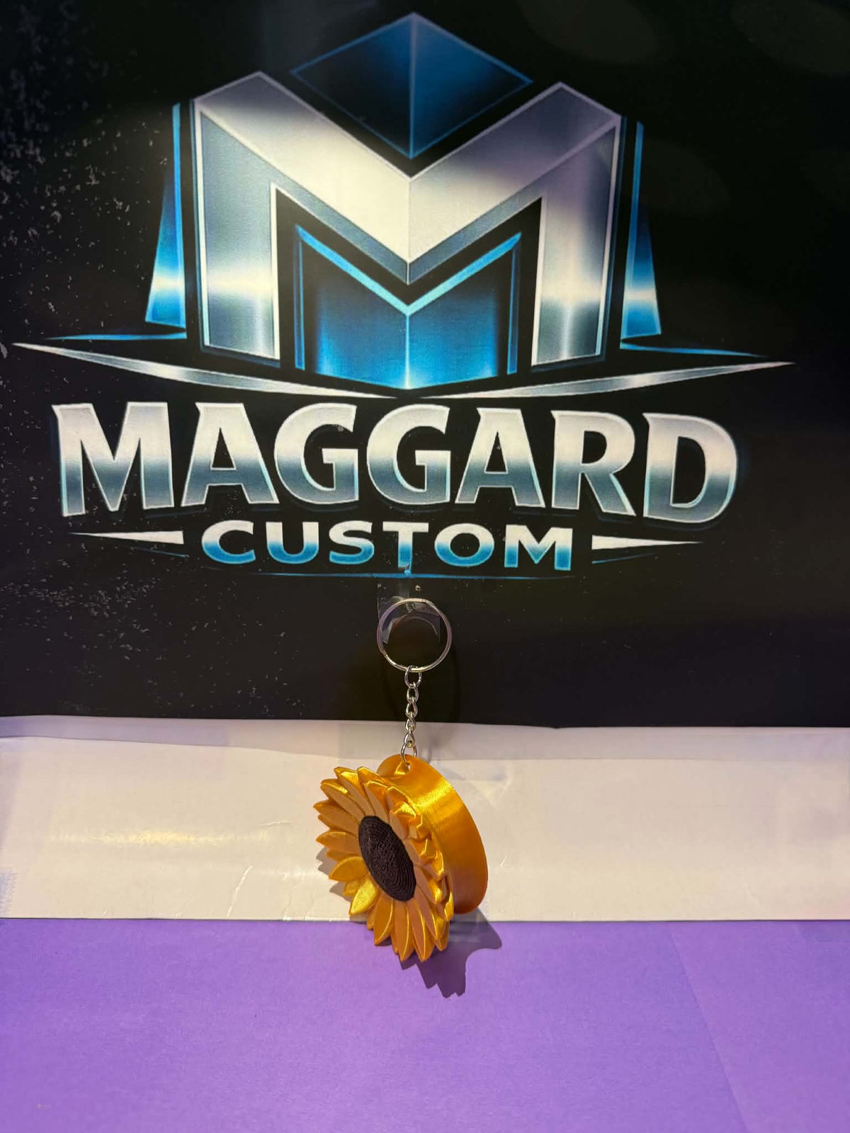 3D printed golden yellow sunflower keychain hair tie holder with layered petals and textured seed center on metal keyring displayed in front of Maggard Custom logo