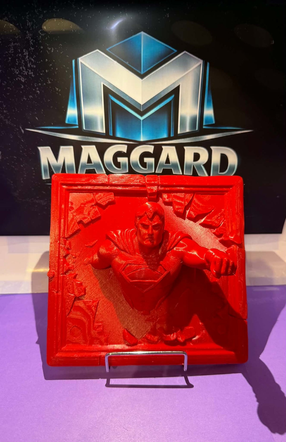 3D printed red caped hero breaking through wall bas-relief plaque with crumbling stone detail displayed on easel in front of Maggard Custom logo
