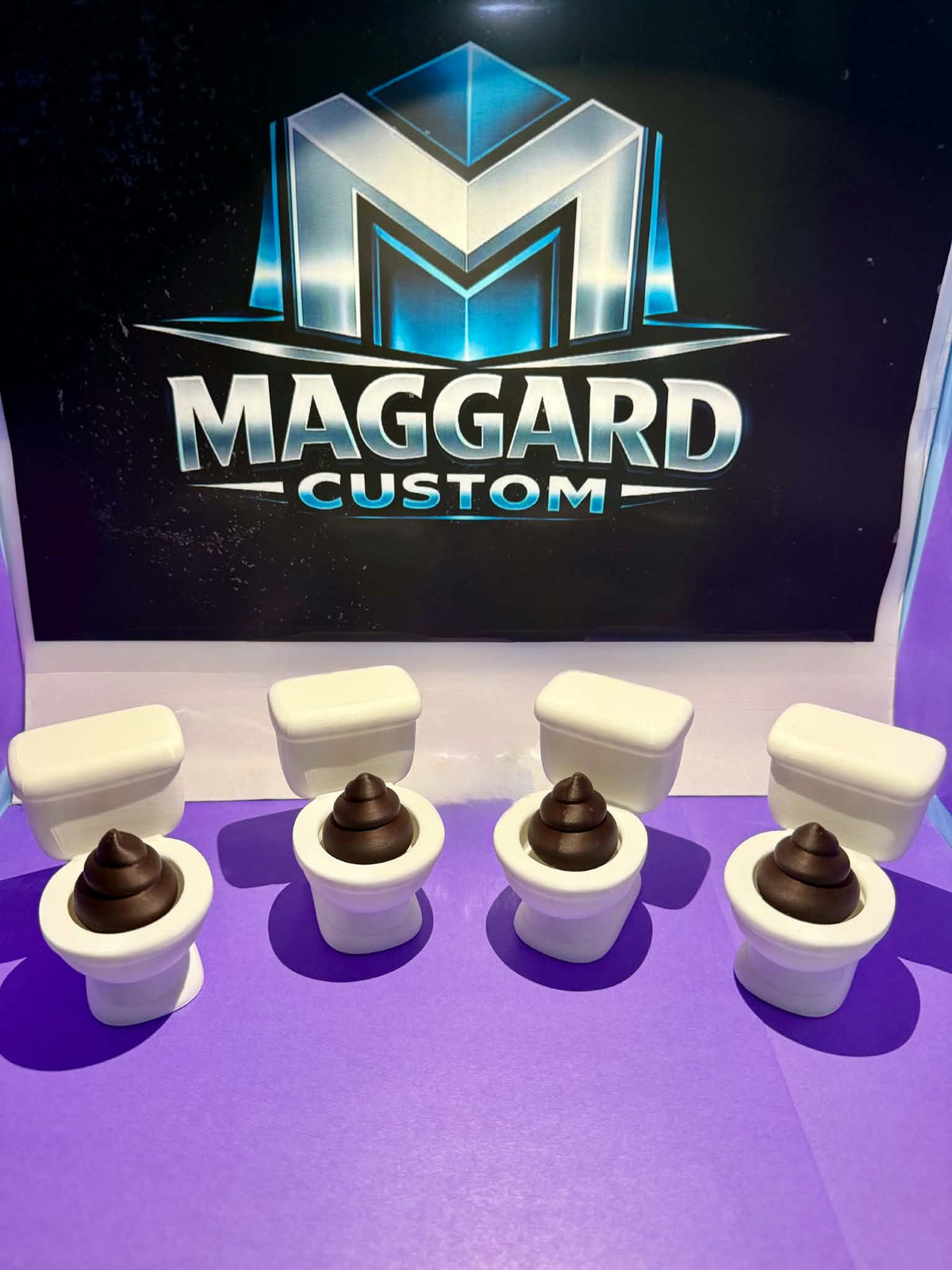 3D printed white toilet bowl fidget clickers with brown surprise inside displayed in front of Maggard Custom logo