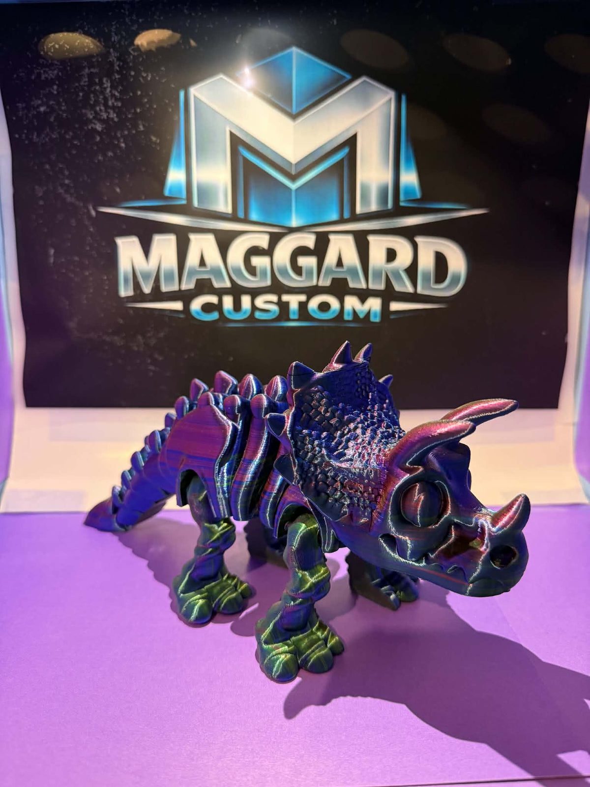 3D printed articulated triceratops figurine with iridescent galaxy finish shifting purple, green, and blue displayed in front of Maggard Custom logo